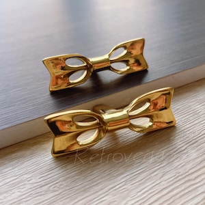 May include: Two gold-colored bow-shaped decorative objects. Each bow has a central knot and two loops with cut-out details. The objects are displayed on a light-colored wooden surface with a dark wooden edge.