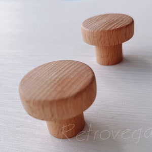 Può includere: Due pomelli in legno con la parte superiore rotonda e una base cilindrica. I pomelli sono in legno marrone chiaro con una venatura naturale.