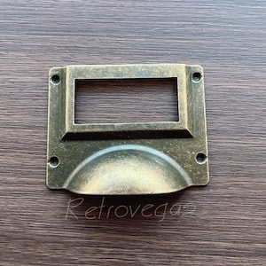 Puede incluir: Un tirador de cajón de latón con un portaetiquetas rectangular y una manija redondeada ligeramente hundida. El tirador tiene un aspecto vintage y antiguo.
