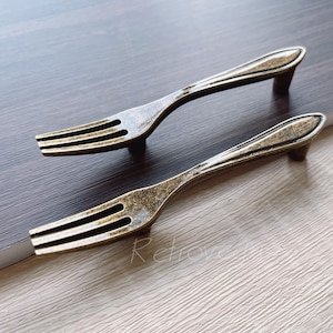 Könnte beinhalten: Zwei antike goldfarbene Kabinettgriffe in Form einer Gabel. Die Griffe haben einen Vintage-Look und sind perfekt, um jedem Wohndesign einen Hauch von rustikalem Charme zu verleihen.