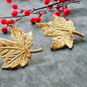 Może przedstawiać: Dwa złote kolczyki w kształcie liścia klonu. Liście są szczegółowe i mają błyszczące wykończenie.