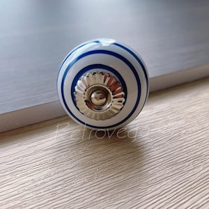 Może przedstawiać: Biały ceramiczny gałka z niebieskimi paskami i srebrnym środkiem. Gałka ma ozdobny wzór wokół środka.