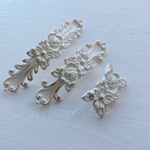 May include: Three white metal decorative drawer pulls with a floral design and gold accents. The pulls are ornate and have a vintage style.