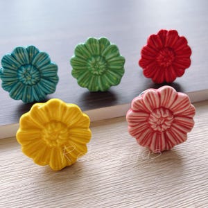 Puede incluir: Cinco tiradores de cajones de cerámica con forma de flor en varios colores. Los tiradores son turquesa, verde, rojo, rosa y amarillo.