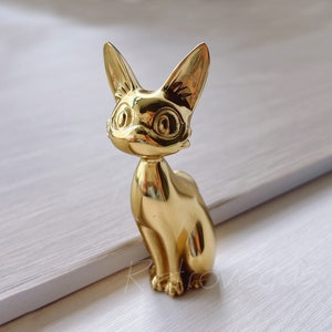 May include: A gold-coloured metal figurine of a cat sitting with its ears perked up. The cat has large, round eyes and a detailed face.