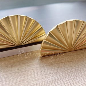 Può includere: Due tiretti per cassetti in metallo color oro a forma di ventaglio. I tiretti sono semicircoli con un design a forma di sole.