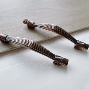 Może przedstawiać: Dwa uchwyty do szafek w kolorze brązu z żebrowanym wzorem. Uchwyty mają zakrzywiony kształt i są wykonane z metalu. Końce uchwytów mają owinięty wzór. Te dekoracyjne uchwyty nadają się do renowacji mebli lub nowych konstrukcji.