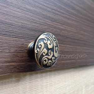 Può includere: Primo piano di un pomello decorativo per mobili con un design a volute in oro antico su uno sfondo scuro e strutturato. Il pomello ha una base nera e una forma rotonda, per un tocco vintage.