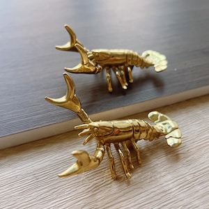 May include: Two gold-colored lobster figurines. The lobsters are detailed with claws, legs, and segmented bodies. They are positioned on a wooden surface, with one slightly above the other. The figurines have a shiny, metallic finish.