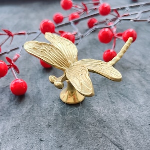 May include: Gold dragonfly door knob with a detailed design. The knob is on a grey fabric background.