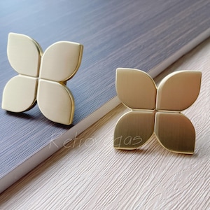 May include: Two gold-toned metal drawer pulls in the shape of a four-leaf clover. The pulls have a brushed finish.