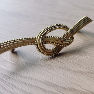 May include: A gold-toned metal cabinet pull with a knot design. The pull has a rope-like texture and is designed to be used on cabinets or drawers.