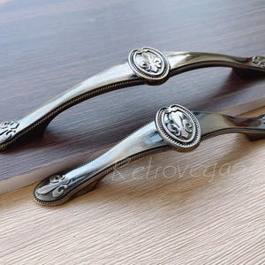 May include: Two antique-style cabinet pulls with a fleur-de-lis design. The pulls are made of metal and have a brushed finish.