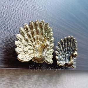 May include: Two gold peacock-shaped drawer pulls with detailed feather designs. The pull on the left is gold, and the pull on the right is a mix of gold and silver.