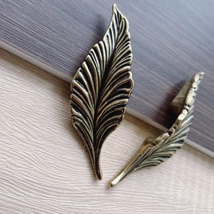 May include: Two gold-toned leaf-shaped cabinet pulls with a detailed, textured design.