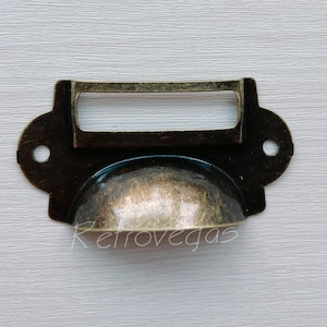 Puede incluir: Tirador de cajón de latón envejecido con asa curva y portaetiquetas rectangular. El herraje tiene un acabado envejecido y dos orificios para tornillos. Ideal para muebles antiguos.