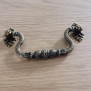 May include: A decorative antique-style cabinet pull with a serpentine design and ornate details. The pull is made of metal with a brass finish.