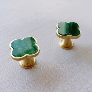May include: Two gold cabinet knobs with a green clover-shaped design. The knobs have a smooth, polished finish.