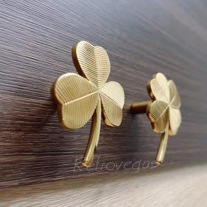 May include: Two gold-toned metal cabinet knobs shaped like four-leaf clovers. The knobs are mounted on a dark brown wooden surface.