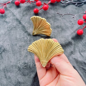 Peut inclure: Deux poignées de tiroir en métal doré en forme de feuille de ginkgo. Les poignées sont présentées sur un fond en tissu gris.