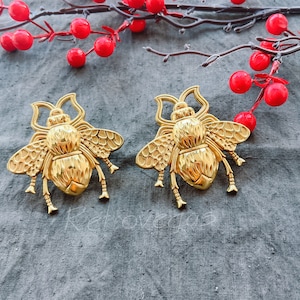 Może przedstawiać: Dwa złote broszki w kształcie pszczoły z detalicznymi skrzydłami i fakturowanymi korpusami. Broszki znajdują się na szarym tle z tkaniny.