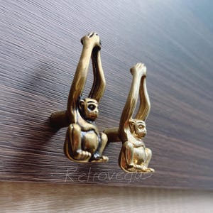 May include: Two gold monkey drawer pulls with arms raised above their heads. The monkey's faces are detailed and the pulls are made of metal.