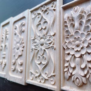 May include: Four rectangular wooden panels with intricate floral and scrollwork carvings. The panels are light-colored, possibly unfinished, and arranged in a row, showcasing detailed relief patterns. The carvings include flowers, leaves, and swirling designs.