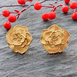 May include: A pair of gold-tone flower-shaped earrings. The earrings are made of metal and have a layered design.
