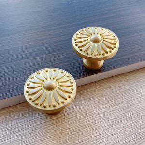 May include: Two gold-toned cabinet knobs with a circular, floral design. The knobs have a raised, textured surface and a smooth, round base.
