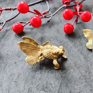 May include: A gold-coloured metal figurine of a goldfish. The figurine is detailed with scales and fins. The goldfish is sitting on a grey textured surface.