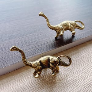 Puede incluir: Dos tiradores de cajón en forma de dinosaurio de color dorado. Los dinosaurios están de pie con la cola enroscada.