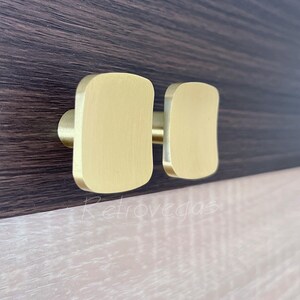 Puede incluir: Un par de tiradores dorados para armario con diseño rectangular. Montados sobre una superficie de madera marrón oscuro, con un panel de madera más claro debajo. La ferretería tiene un acabado liso y mate.