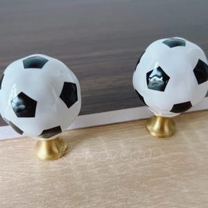 May include: Two cabinet knobs featuring a football design. The knobs are white with black pentagons, mounted on a gold-coloured base. They are set against a light brown wooden surface.