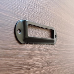 May include: Black metal label holder with a rectangular opening for inserting a label. The holder has rounded edges and a smooth finish.