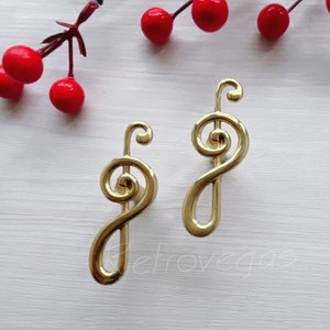 May include: Two gold-colored, stylized treble clef cabinet pulls. The pulls have a curved design with a spiral at the top and a long, slender stem. Red berries and a sprig of greenery are in the background.