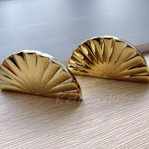 May include: Two gold-toned metal cabinet pulls shaped like half a sunburst. The pulls have a fan-like design with a smooth, shiny finish.