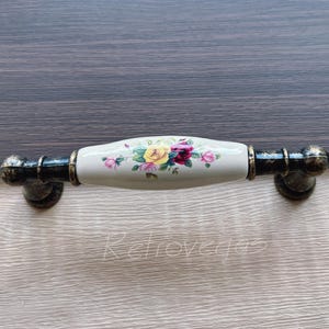 Könnte beinhalten: Ein weißer Keramik-Schubladengriff mit einem Blumendesign in Rosa-, Gelb- und Grüntönen. Der Griff hat schwarze Metallenden.