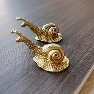 May include: Two gold-colored snail-shaped decorative objects. Each snail has a textured body and a spiraled shell. The snails are positioned on a dark brown wooden surface.