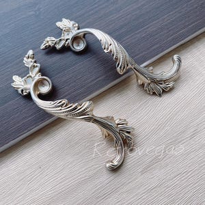 Op de afbeelding: Twee sierlijke, goudkleurige kastladegrepen met een bladontwerp.