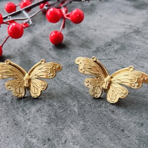 May include: Two gold-colored butterfly-shaped metal charms. The butterflies have detailed wings and a textured surface.