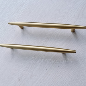 May include: Two gold-toned metal cabinet pulls with a long, thin, and slightly tapered design. The pulls are arranged horizontally, one above the other.