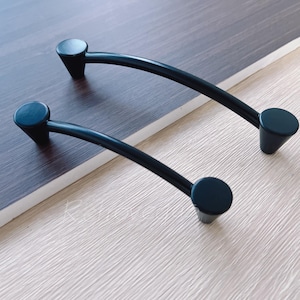 May include: Two black metal cabinet pulls with a curved design and rounded ends. The pulls are shown on a wooden surface.