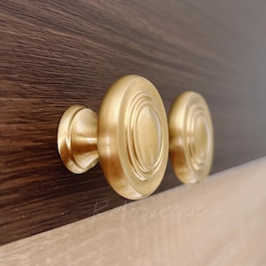 May include: Close-up of two gold-colored cabinet knobs with a circular design. The knobs are mounted on a dark brown wooden surface. The knobs have a classic, elegant appearance.