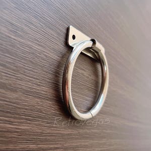 May include: Silver metal ring handle with a rectangular mounting plate. The handle is attached to a wooden surface.
