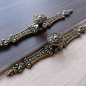 May include: Two ornate gold-toned cabinet drawer pulls with intricate floral designs. Each pull features a round knob with a raised floral pattern.