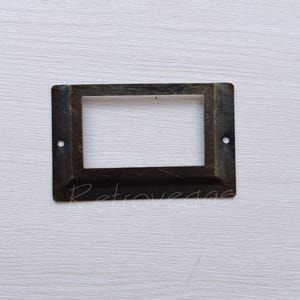 May include: A brass drawer label holder with a rectangular opening. The holder has two small holes on the sides and a slightly raised edge around the opening.