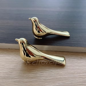 May include: Two gold-coloured ceramic bird figurines. The birds are in a simple, modern style with a smooth, glossy finish.