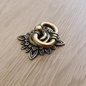May include: A gold-toned metal drawer pull with a decorative floral design. The pull has a ring-shaped handle.