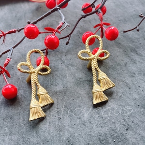 May include: Two gold-toned decorative drawer pulls with a rope design and tassel accents. The pulls are against a grey background with red berries.