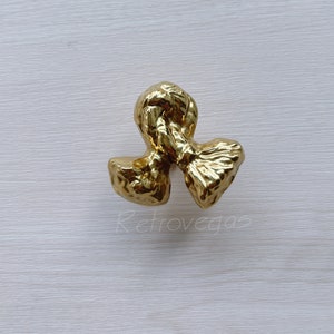 May include: A gold-colored, abstract decorative object with a textured surface. The design features a knot-like central element with two flared, rounded ends. The object is set against a light, neutral background.
